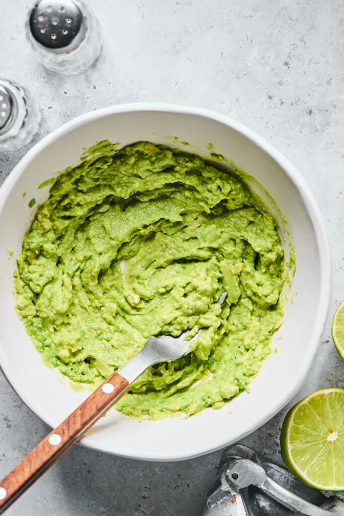 A white bowl filled with guacamole with a fork in it. Around the bowl is a salt and pepper shaker, a lime that has been cut in half, and a silver citrus press.