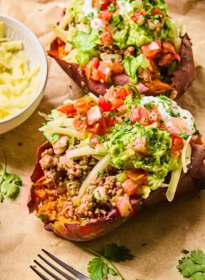 Two taco stuffed sweet potatoes piece of parchment paper. They are stuffed with taco meat, shredded cheese, shredded lettuce, pico de gallo, guacamole, greek yogurt, and cilantro. Around them is a fork, some cilantro leaves, and a bowl of shredded cheese.