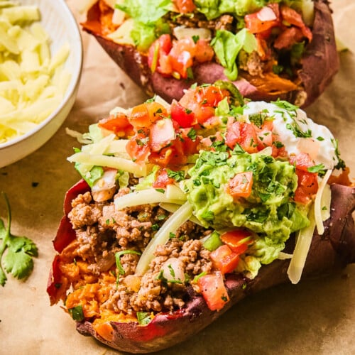Two taco stuffed sweet potatoes piece of parchment paper. They are stuffed with taco meat, shredded cheese, shredded lettuce, pico de gallo, guacamole, greek yogurt, and cilantro. Around them is a fork, some cilantro leaves, and a bowl of shredded cheese.