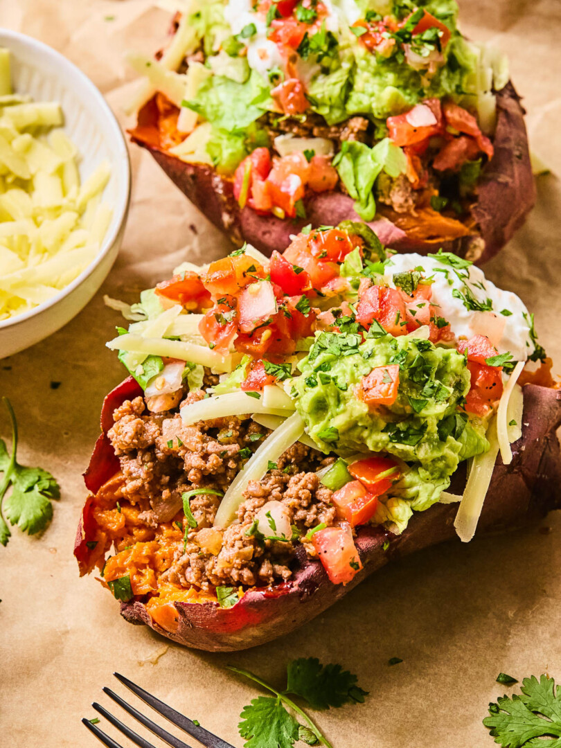 Two taco stuffed sweet potatoes piece of parchment paper. They are stuffed with taco meat, shredded cheese, shredded lettuce, pico de gallo, guacamole, greek yogurt, and cilantro. Around them is a fork, some cilantro leaves, and a bowl of shredded cheese.