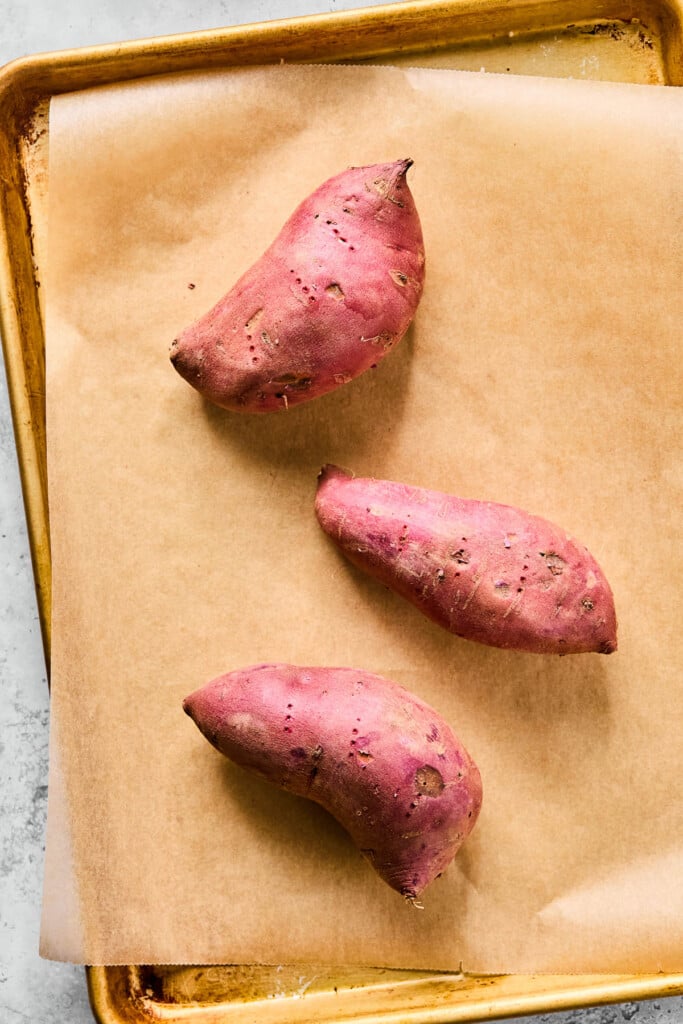 Three uncooked sweet potatoes on a baking sheet lined with parchment paper.