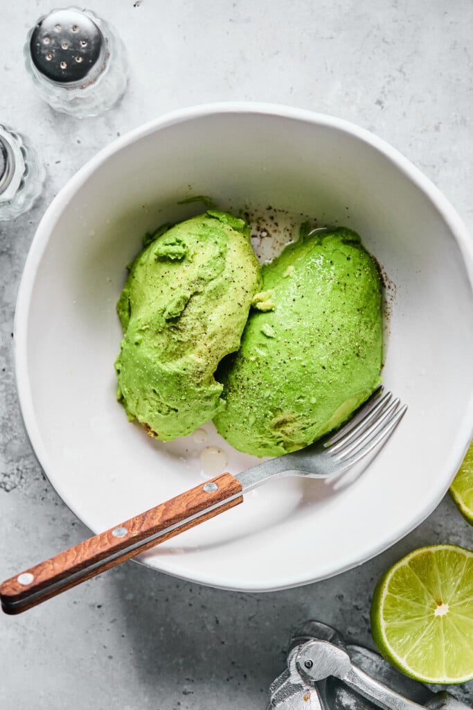 A white bowl with a whole avocado that has been cut in half in it. A fork is in the bowl as well. Around the bowl is a salt and pepper shaker, a lime that has been cut in half, and a silver citrus press.