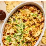 BBQ chicken dip topped with melted cheese and garnished with sliced green onions, cilantro, and BBQ sauce in a white baking dish. Some tortillas chips are in the dip as well. Around the dish are some tortilla chips, a plate with dip on it, and a cup of bbq sauce.
