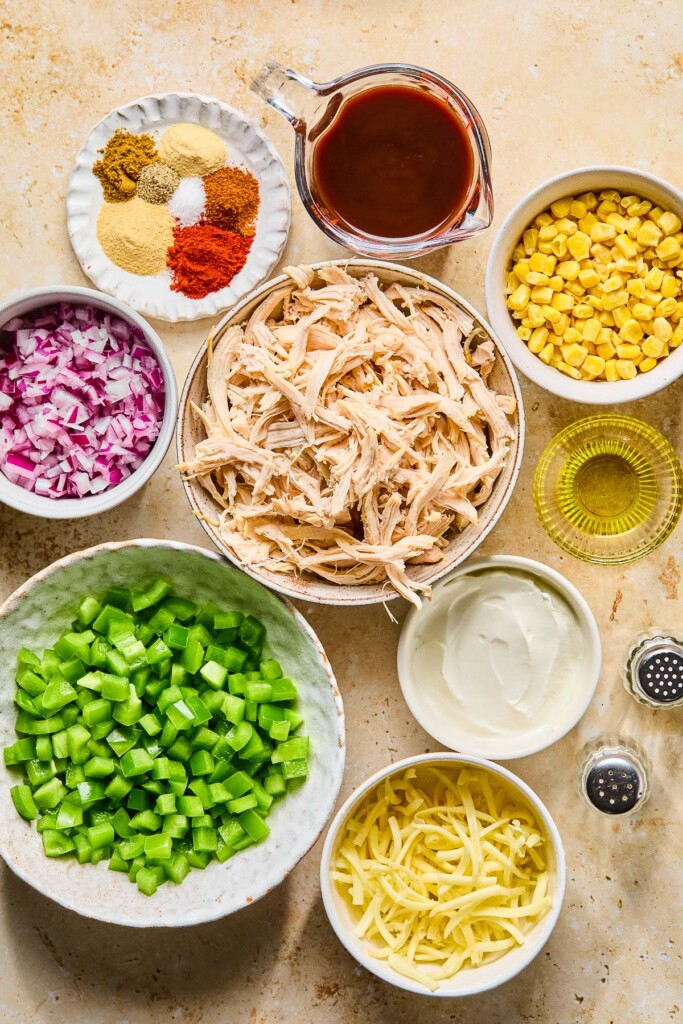 A measuring cup of bbq sauce, a bowl of frozen corn, a small cup of olive oil, a bowl of greek yogurt, a salt and pepper shaker, a bowl of shredded cheese, a bowl of diced green onions, a bowl of shredded chicken, a bowl of diced red onion, and a plate of different piles of spices on a tan counter.