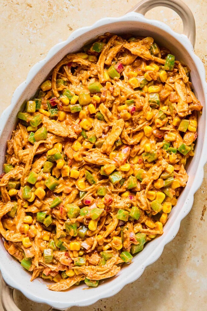 A mixture of shredded chicken and veggies in a creamy brownish sauce in a white au gratin dish before it is baked.