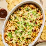 Au gratin dish filled with BBQ chicken dip topped with melted cheese and garnished with sliced green onions, cilantro, and BBQ sauce. Around the dish are some tortilla chips, an empty plate, a cup of bbq sauce, and a plate of cilantro.