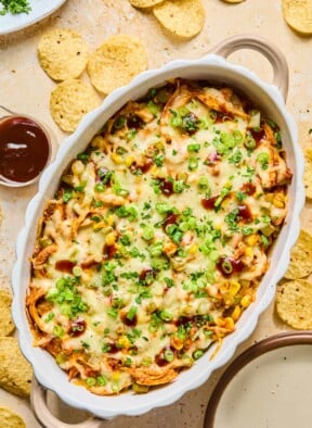 Au gratin dish filled with BBQ chicken dip topped with melted cheese and garnished with sliced green onions, cilantro, and BBQ sauce. Around the dish are some tortilla chips, an empty plate, a cup of bbq sauce, and a plate of cilantro.
