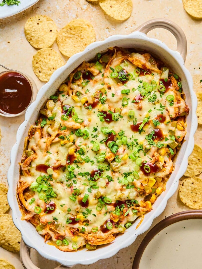Au gratin dish filled with BBQ chicken dip topped with melted cheese and garnished with sliced green onions, cilantro, and BBQ sauce. Around the dish are some tortilla chips, an empty plate, a cup of bbq sauce, and a plate of cilantro.