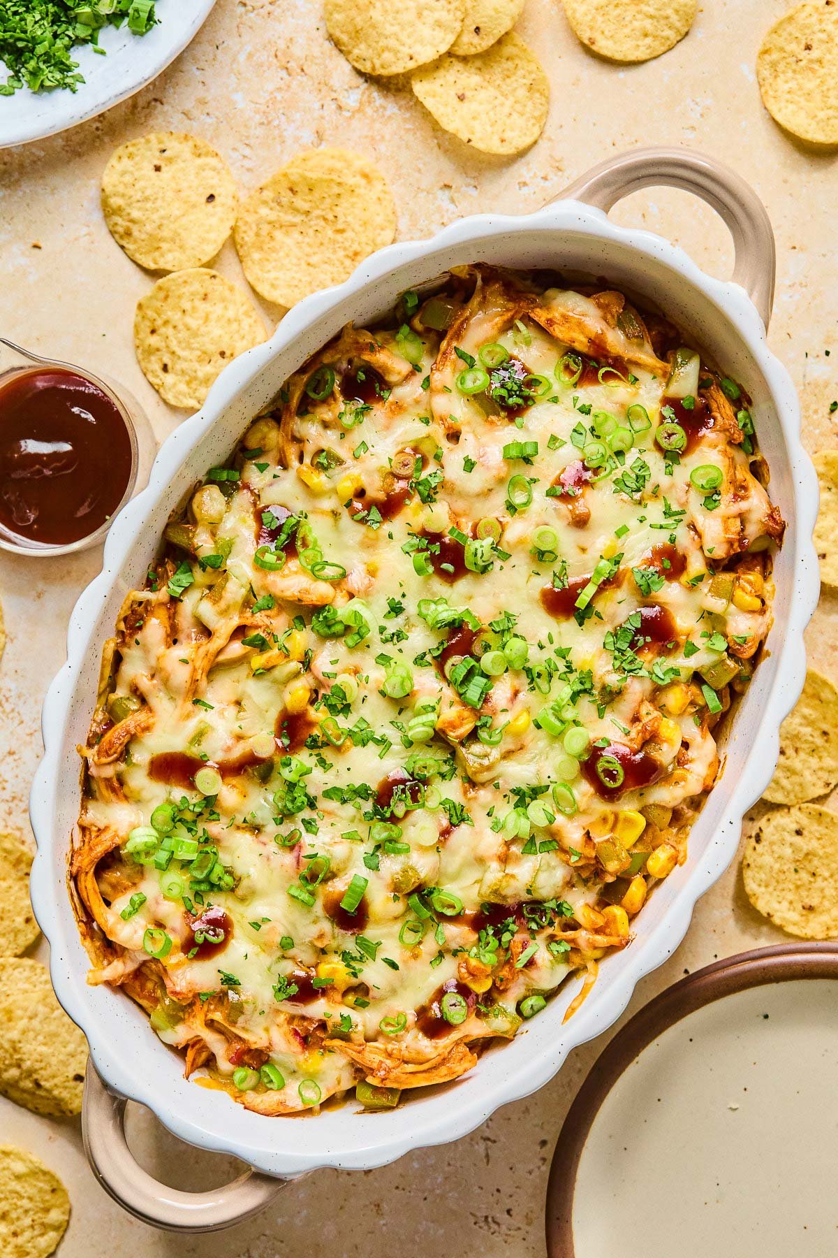 Au gratin dish filled with BBQ chicken dip topped with melted cheese and garnished with sliced green onions, cilantro, and BBQ sauce. Around the dish are some tortilla chips, an empty plate, a cup of bbq sauce, and a plate of cilantro.