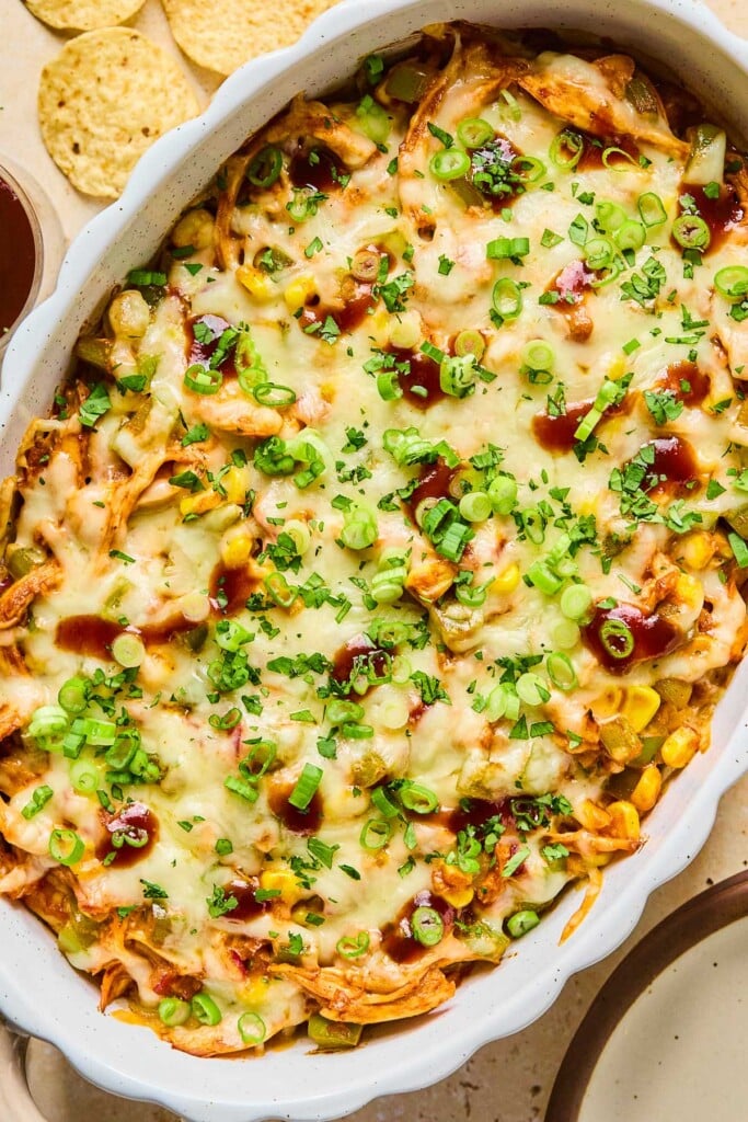 A dish of barbecue chicken dip with corn, veggies, and topped with melted cheese. It is garnished with sliced green onions, cilantro, and BBQ sauce. Around t the pan is an empty plate, some bbq sauce, and tortilla chips.