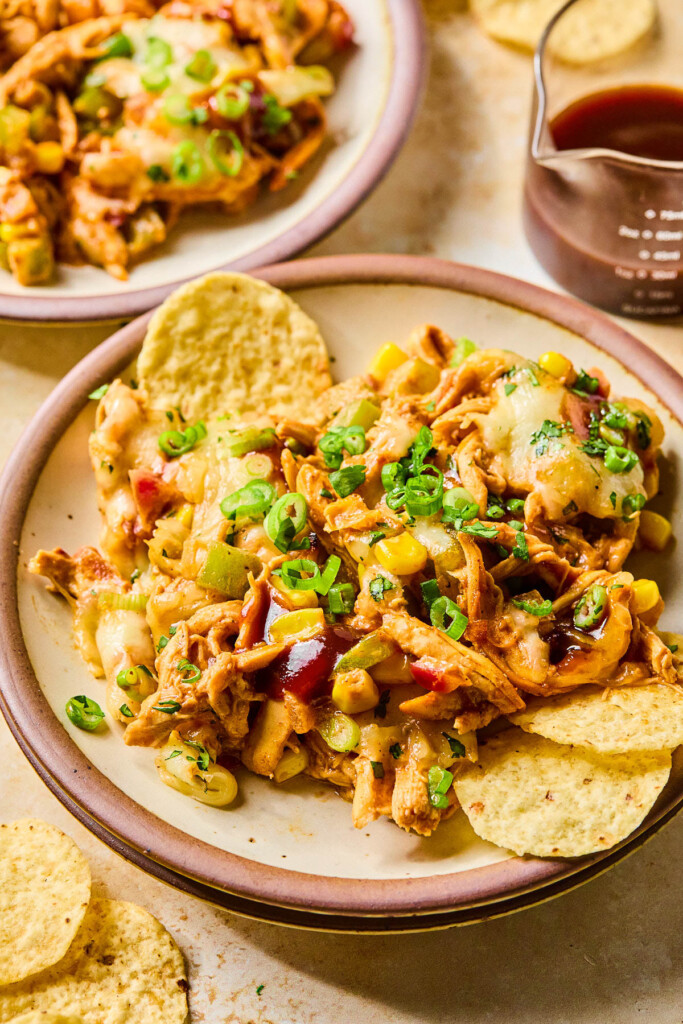 Cheesy BBQ chicken dip on a plate garnished with green onions, cilantro, and bbq sauce. Also on the plate are some tortilla chips. Around the plate are more tortilla chips, another plate of dip, and a measuring cup of bbq sauce.