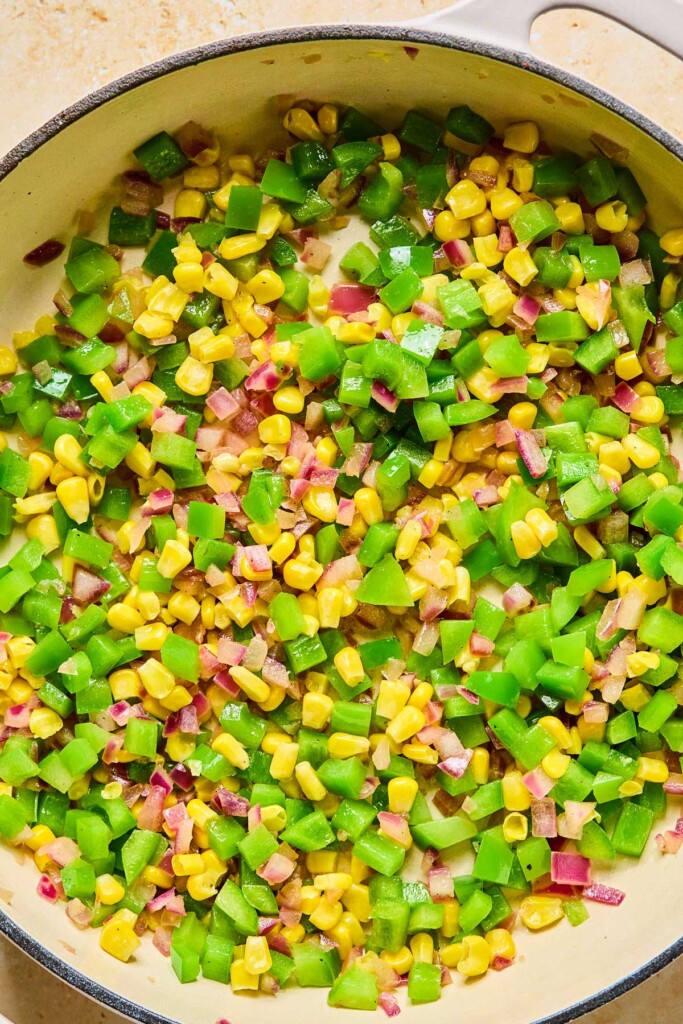 Large enameled pan filled with sautéed red onions, diced green bell peppers, and corn.