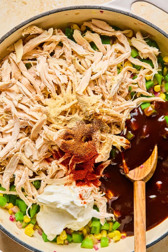 Large enameled pan filled with diced sautéed veggies, shredded chicken, spices, greek yogurt, and BBQ sauce before it is mixed together. A wooden spoon is in the pan as well.