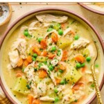 A bowl with healthy chicken pot pie soup with potatoes and veggies and garnished with fresh parsley and black pepper. A spoon is also in the bowl. Around the bowl is a mustard colored kitchen towel and a small bowl of fresh parsley.