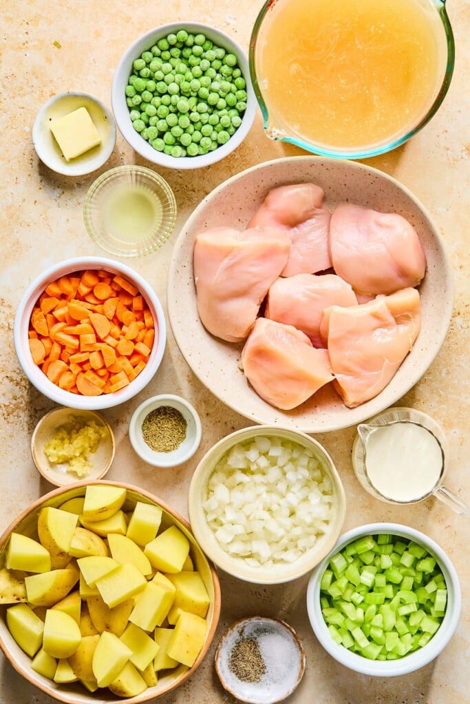 A large measuring cup of broth, a bowl of raw chicken breasts, a small measuring cup of heavy cream, a bowl of diced yellow onions, a bowl of diced celery, a small bowl of salt and pepper, a bowl of diced yukon gold potatoes, a small bowl of minced garlic, a small bowl of dried thyme, a bowl of diced carrots, a small bowl of lemon juice, a small dish with a tablespoon of button on it, and a bowl of frozen peas on a tan counter.
