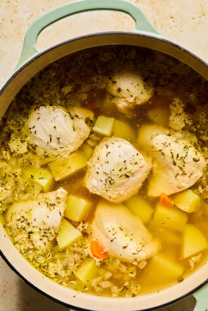 Large dutch oven filled with large pieces of cooked chicken, diced potatoes, diced veggies, and spices in broth.