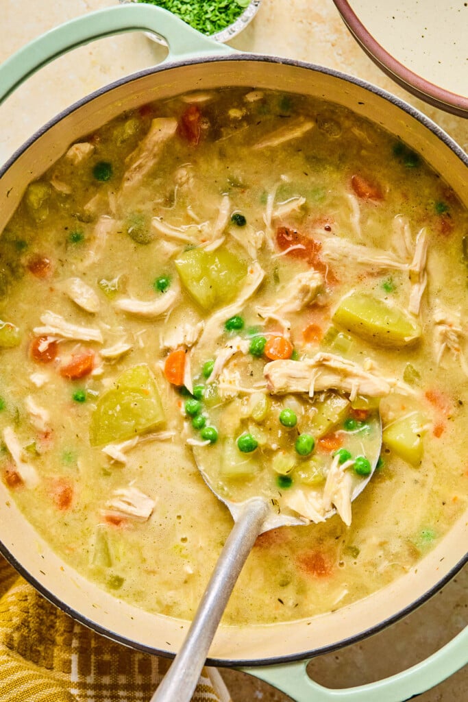 Large dutch oven of chicken pot pie soup with a large ladle in it. Next to the pot is a mustard colored kitchen towel, a bowl of chopped parsley, and an empty bowl.