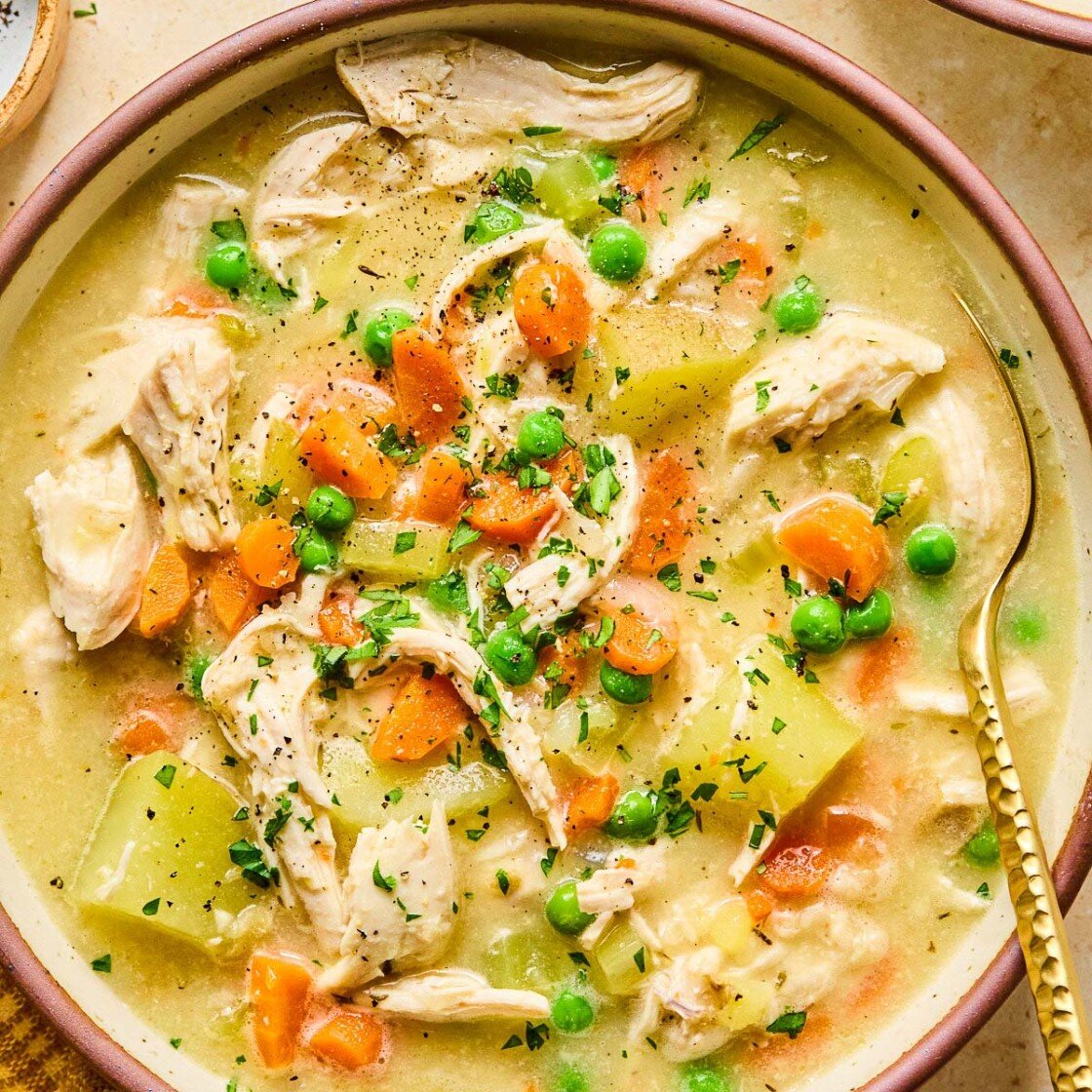 A bowl with healthy chicken pot pie soup with potatoes and veggies and garnished with fresh parsley and black pepper. A spoon is also in the bowl. Around the bowl is a mustard colored kitchen towel and another bowl of soup.