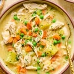 A bowl with healthy chicken pot pie soup with potatoes and veggies and garnished with fresh parsley and black pepper. A spoon is also in the bowl. Around the bowl is a mustard colored kitchen towel and another bowl of soup.