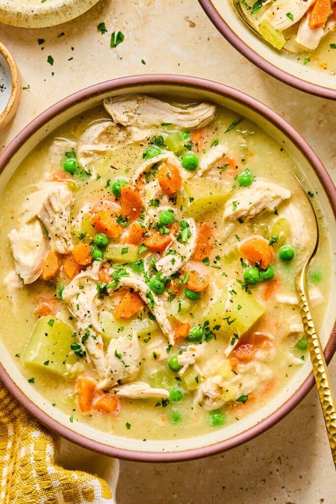 A bowl with healthy chicken pot pie soup with potatoes and veggies and garnished with fresh parsley and black pepper. A spoon is also in the bowl. Around the bowl is a mustard colored kitchen towel and another bowl of soup.