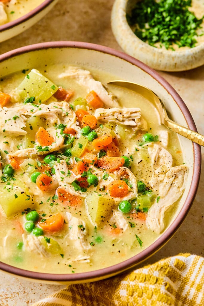 A bowl of creamy chicken pot pie soup with potatoes and garnished with fresh parsley. A spoon is also in the bowl. Around the bowl is a mustard colored kitchen towel, a small bowl of chopped parsley, and another bowl of soup.