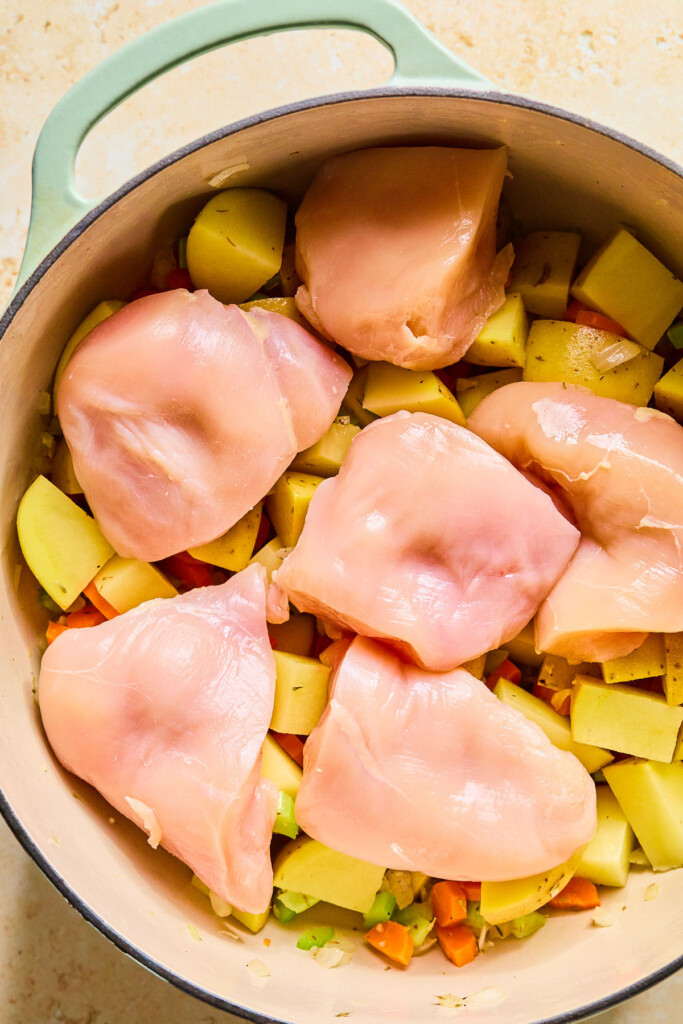 Large dutch oven filled with a mix of sautéed veggies and raw diced potatoes with large pieces of raw chicken breasts sitting on top of the mix.