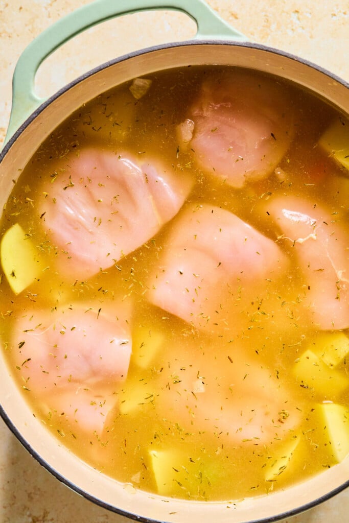 Large dutch oven filled with large pieces of raw chicken, diced potatoes, and dried thyme in broth before it is cooked.