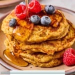 A stack of greek yogurt pancakes topped with raspberries and blueberries on a plate. Maple syrup is being poured on them. Around the plate are more berries and another stack of pancakes.
