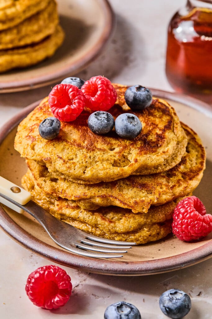 A stack of pancakes with greek yogurt topped with raspberries and blueberries on a plate. A fork is also on the plate. Around the plate are more berries, a glass jar of maple syrup, and another stack of pancakes.