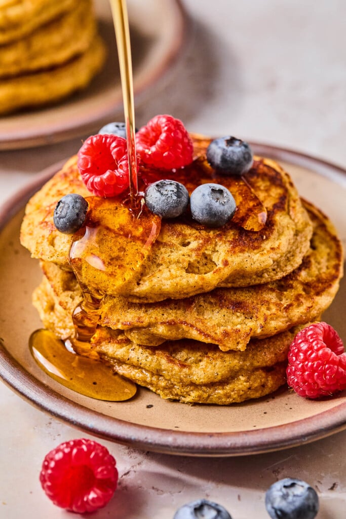 A stack of greek yogurt pancakes topped with raspberries and blueberries on a plate. Maple syrup is being poured on them. Around the plate are more berries.
