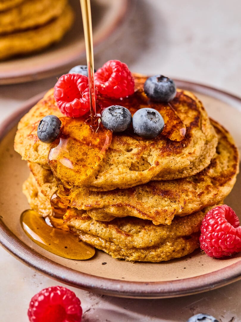 A stack of greek yogurt pancakes topped with raspberries and blueberries on a plate. Maple syrup is being poured on them. Around the plate are more berries.