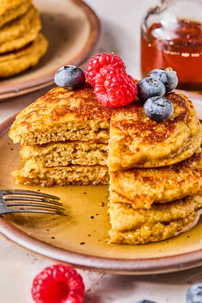 Three yogurt pancakes stacked on top of each other on a plate with a fork resting on it. They are topped with fresh raspberries, blueberries, and maple syrup, and a section is cut out of them. Around the plate are more berries, a glass jar of maple syrup, and another plate with a stack of pancakes.