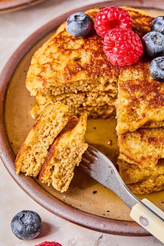 Greek yogurt pancakes stacked on a plate with a section cut out of them on a fork. They are topped with fresh raspberries and blueberries. Around the plate are some more berries.