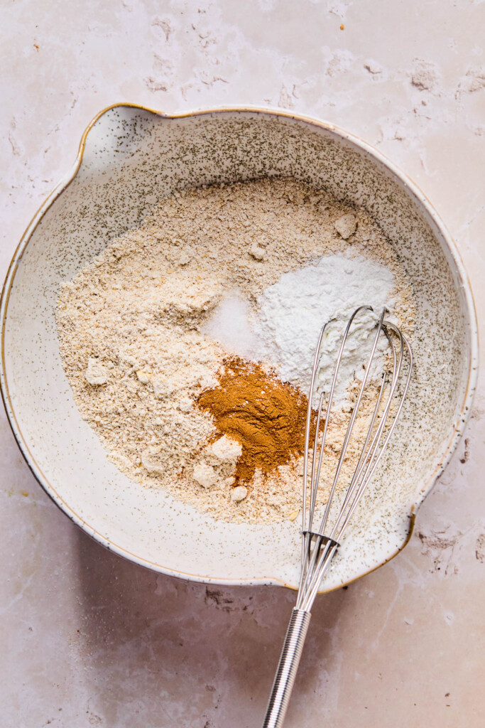 An overhead shot of a bowl with oat flour, cinnamon, baking powder, and salt before it is mixed together. A whisk is also in it.