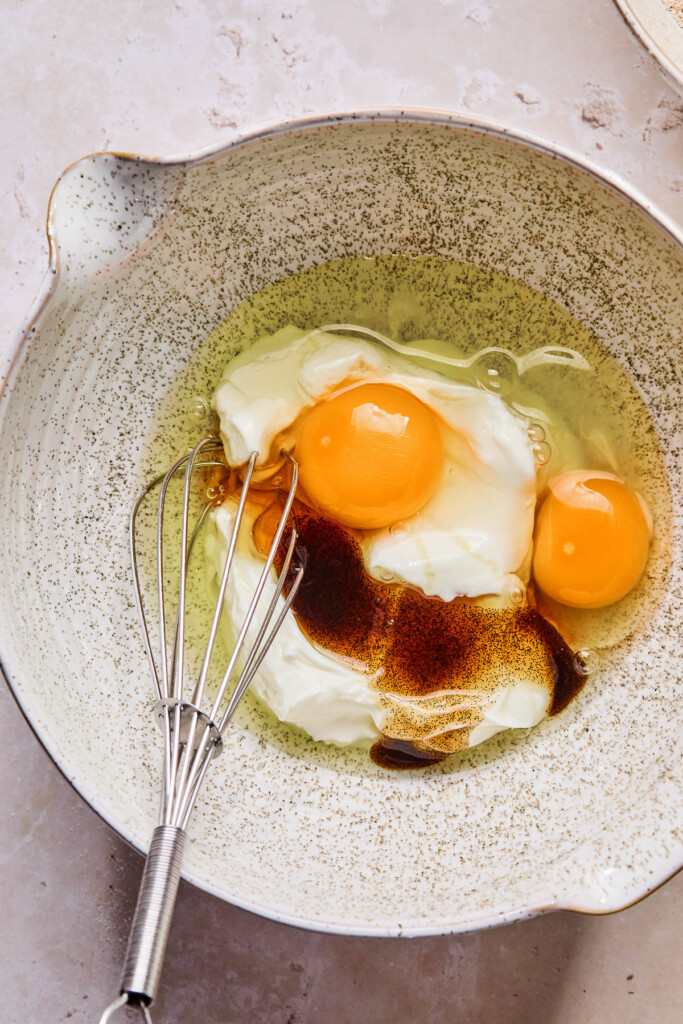 Mixed bowl of greek yogurt, two eggs, honey, and vanilla bean paste before it is mixed together. A whisk is also in the bowl.