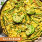 Asparagus frittata with sweet potatoes and goat cheese in a large enameled skillet. It is topped with sliced avocado and fresh parsley. Around the pan is a tan kitchen towel, a small dish of chopped fresh parsley, and another empty plate.