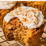 Healthy carrot cake muffin with greek yogurt frosting on a wire cooling rack. It has a bite taken out of it. More carrots cake muffins are behind it.