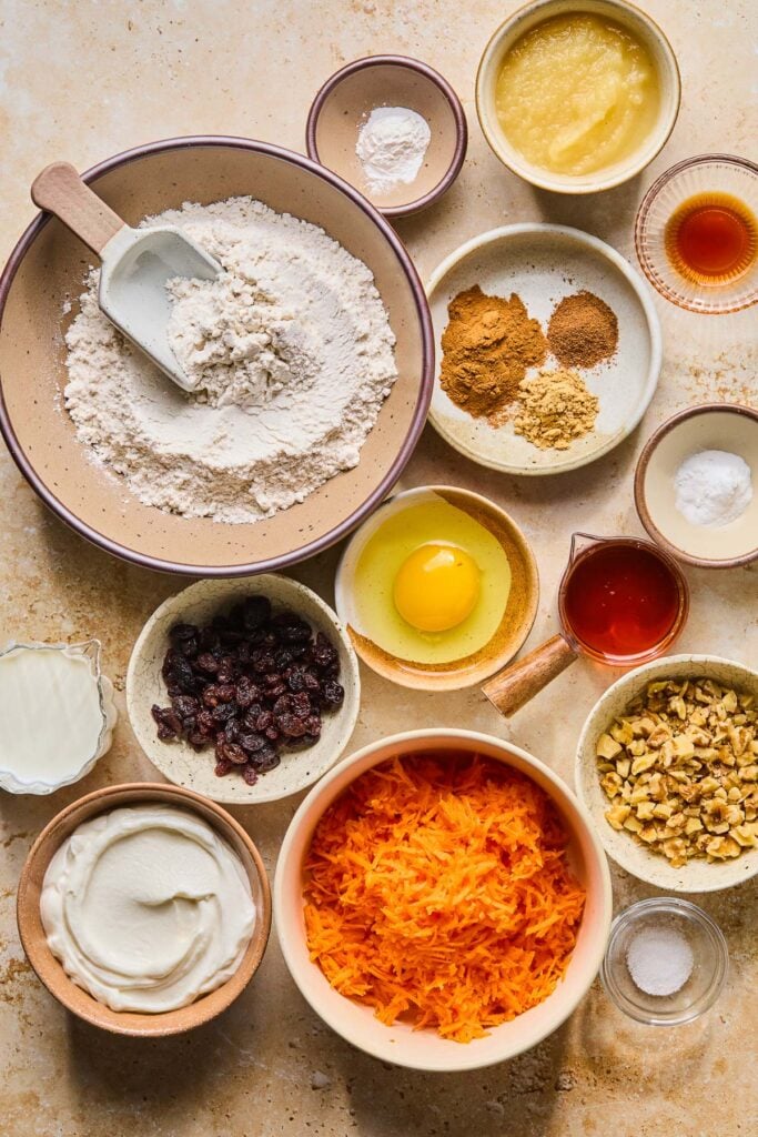 A bowl of applesauce, a small bowl of vanilla extract, a plate of a few piles of different spices, a small bowl of baking powder, a measuring cup of maple syrup, a bowl of chopped walnuts, a small bowl of salt, a bowl of grated carrots, a bowl of greek yogurt, a cup of milk, a bowl of raisins, a bowl with a cracked egg in it, a bowl with flour and a flour scoop in it, and a small bowl of baking soda on a tan counter.