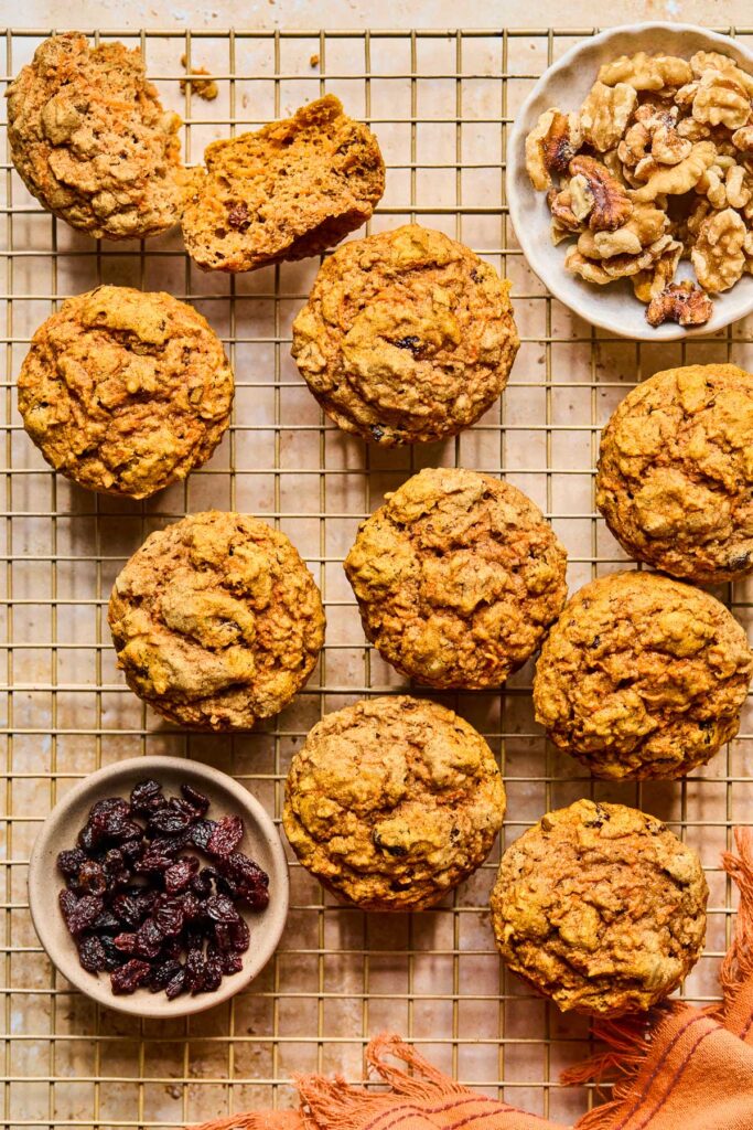 A wire cooling rack with carrots cake muffins spread across it. One of the muffins is broken open. Also on the rack is a bowl of raisins, bowl of walnuts, and an orange kitchen towel.