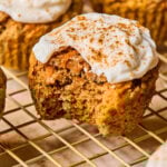 Healthy carrot cake muffins with greek yogurt frosting on a wire cooling rack. The one in front has a bite taken out of it.
