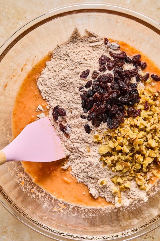 A bowl of wet orange batter with a flour mixture, chopped walnuts, and raisins on top of it before it is mixed. A spatula is also in the bowl.