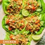 A plate of Asian chicken lettuce wraps with ground chicken, veggies, scallions, peanuts, and sesame seeds. Also on the plate are some lime wedges. Around the plate green kitchen towel, a bowl of sliced green onions, a bowl of cilantro, and a bowl of peanuts.