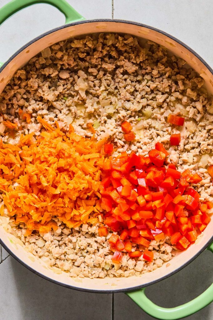 Overhead shot of a large enameled pan filled with sautéed ground chicken with a pile of raw shredded carrots and a pile of diced red bell pepper on top.