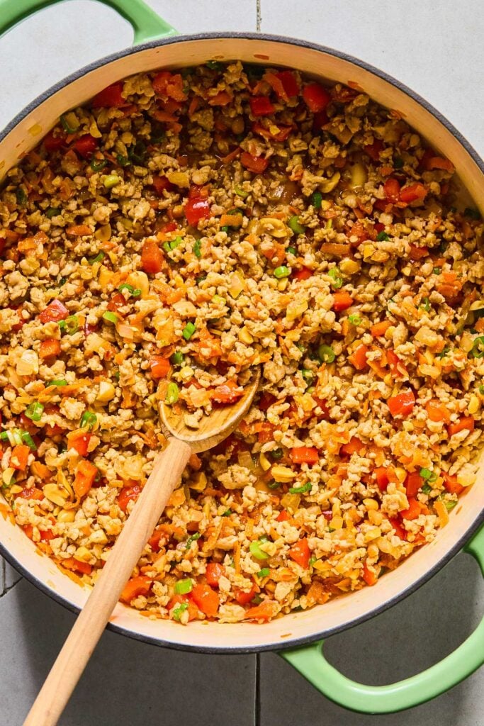 Overhead shot of a large enameled pan filled with sautéed ground chicken, onions, red bell peppers, and shredded carrots, green onions, and peanuts all mixed with a stir fry sauce. A wooden spoon is in the pan as well.