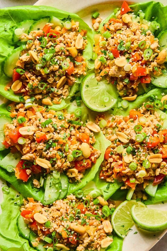 Healthy chicken lettuce wraps with ground chicken, bell peppers, carrots, scallions, and peanuts with cucumbers in lettuce cups. They are garnished with sesame seeds and cilantro. Also on the plate are some lime wedges.