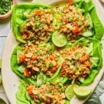 A plate of Asian chicken lettuce wraps with ground chicken, veggies, scallions, peanuts, and sesame seeds. Also on the plate are some lime wedges. Around the plate green kitchen towel, a bowl of sliced green onions, a bowl of cilantro, and a bowl of peanuts.