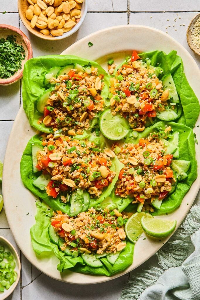 A plate of Asian chicken lettuce wraps with ground chicken, veggies, scallions, peanuts, and sesame seeds. Also on the plate are some lime wedges. Around the plate green kitchen towel, a bowl of sliced green onions, a bowl of cilantro, and a bowl of peanuts.