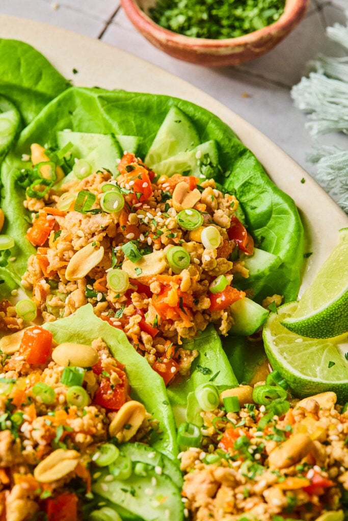 Ground chicken lettuce wraps with veggies, peanuts, and scallions on a plate. They are garnished with sesame seeds and some lime wedges are on the plate. Around the plate is a bowl of chopped cilantro.