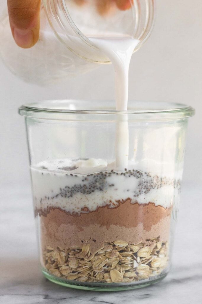 Jar filled with rolled oats, chocolate protein powder, cacao powder, and chia seeds with milk being poured into it.