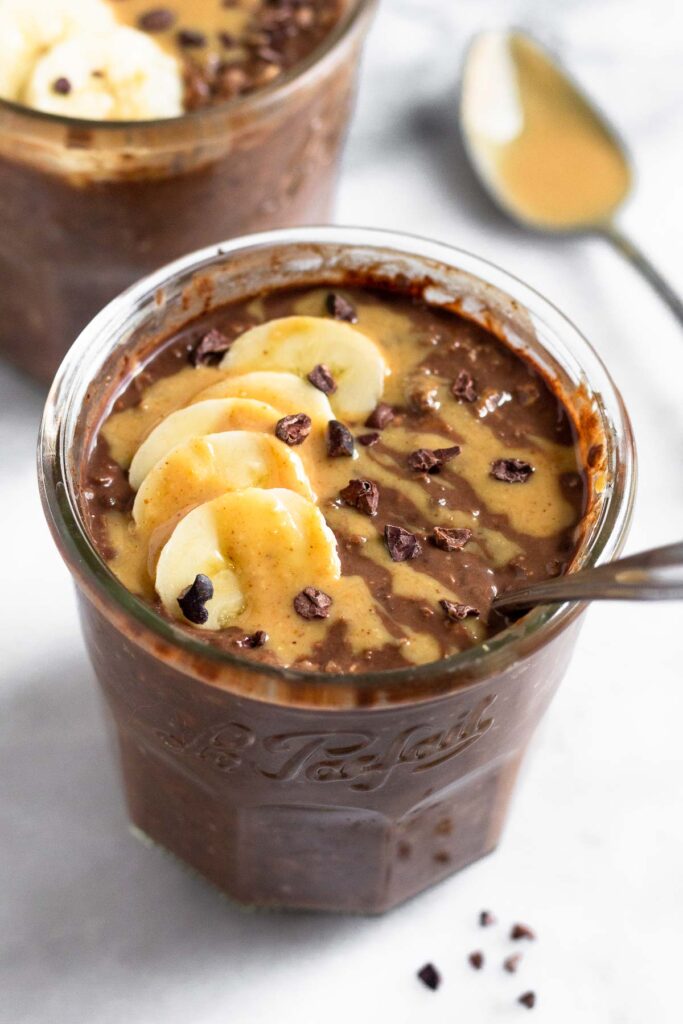Chocolate peanut butter overnight oats in a jar topped with sliced banana, peanut butter, and cacao nibs with a spoon in the jar. Behind it is a another jar of overnight oats and a spoon with peanut butter on it.